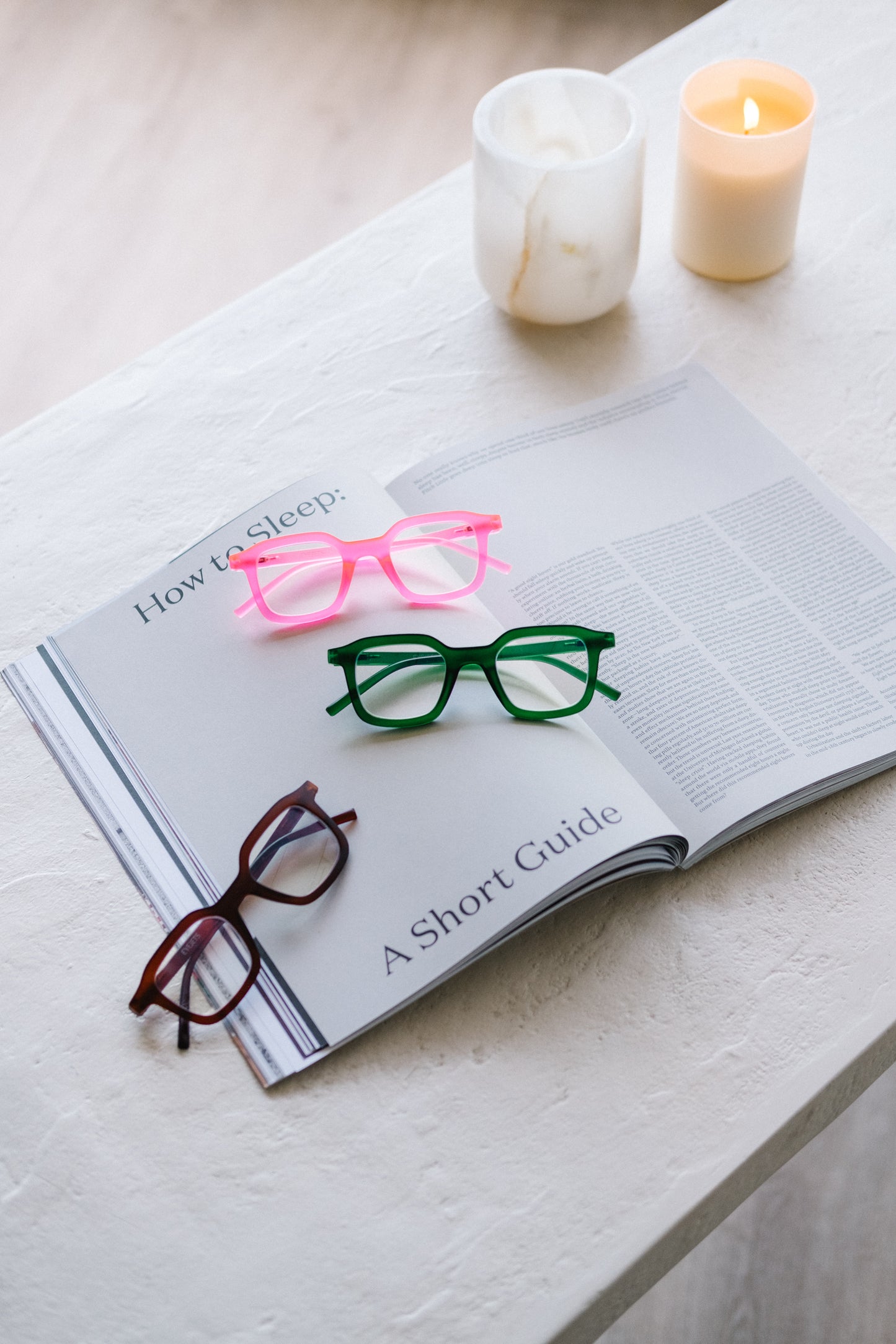 Three Ibiza blue light reading glasses in pink, green, and brown resting on a white desk, placed on top of an open magazine showing an article titled “How to Sleep,” with a lit candle in the corner. The scene conveys chic, relaxing nighttime vibes, highlighting the glasses as stylish accessories that support healthy melatonin production.