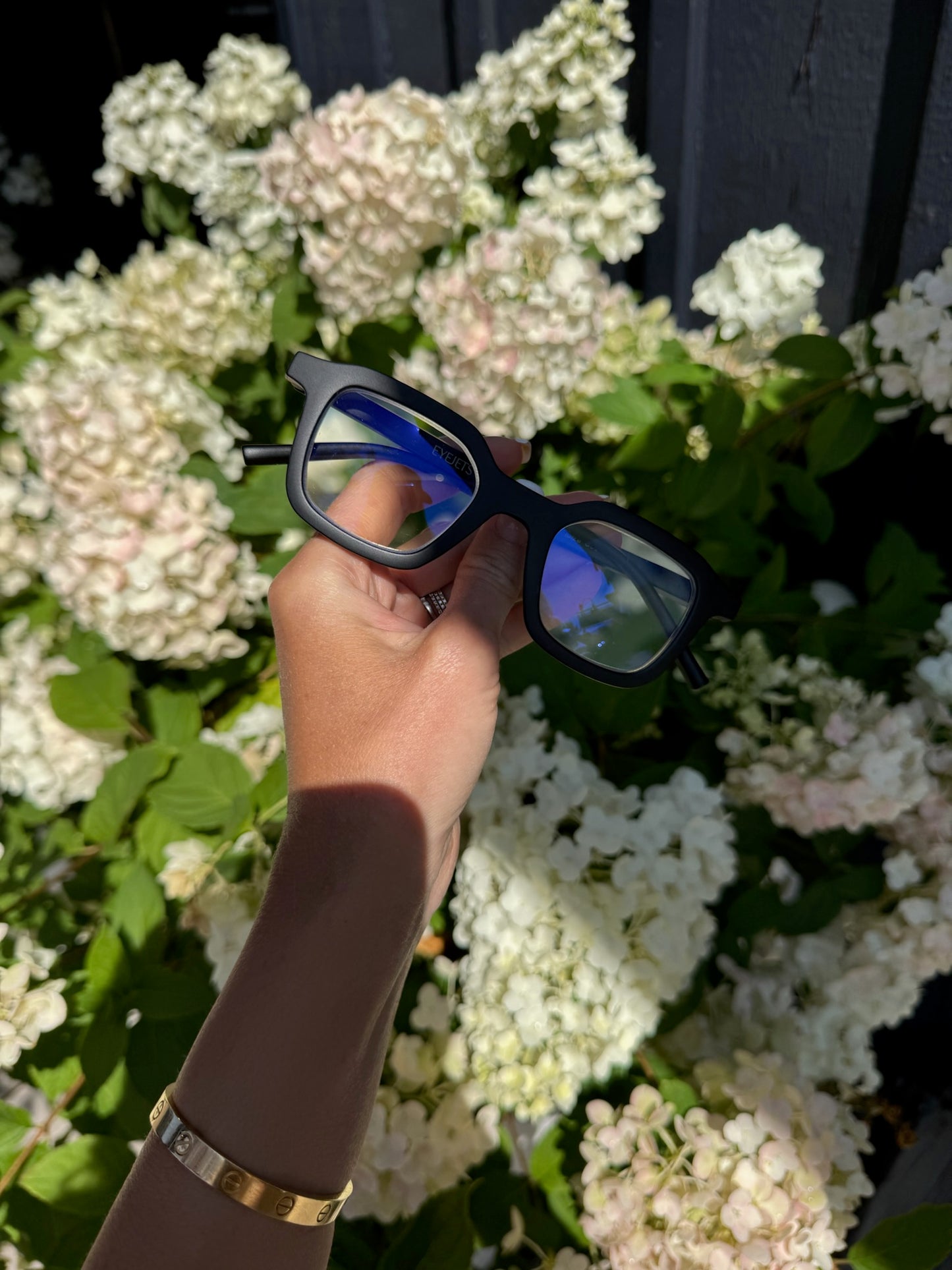 Hand holding matte black Ibiza reading glasses with blue light blocking lenses in front of blooming hydrangea flowers