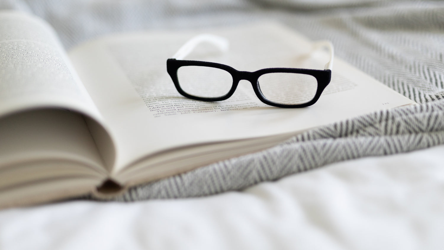 Black and white Sydney velvet reading glasses resting on the pages of an open book on a cozy bed