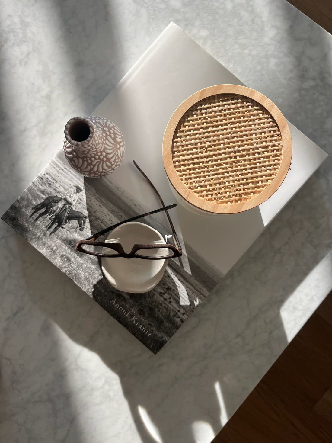A cozy desk setup featuring a pair of reading glasses, an unlit candle, a magazine, and decorative home accessories arranged on a marble coffee table.