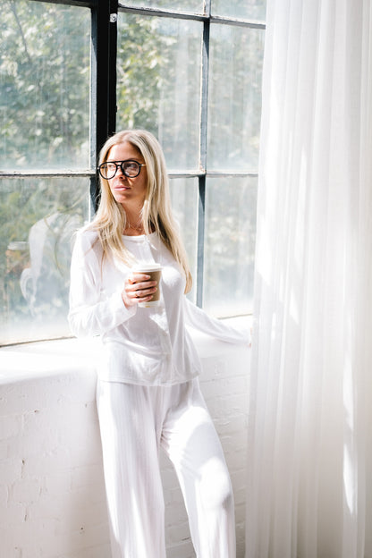 Woman wearing black velvet Mykonos aviator reading glasses, dressed in white pajamas, standing in front of a brightly lit window while holding a cup of coffee and gazing outside. The scene feels calm and cozy, highlighting the elegant velvet frames in natural morning light.