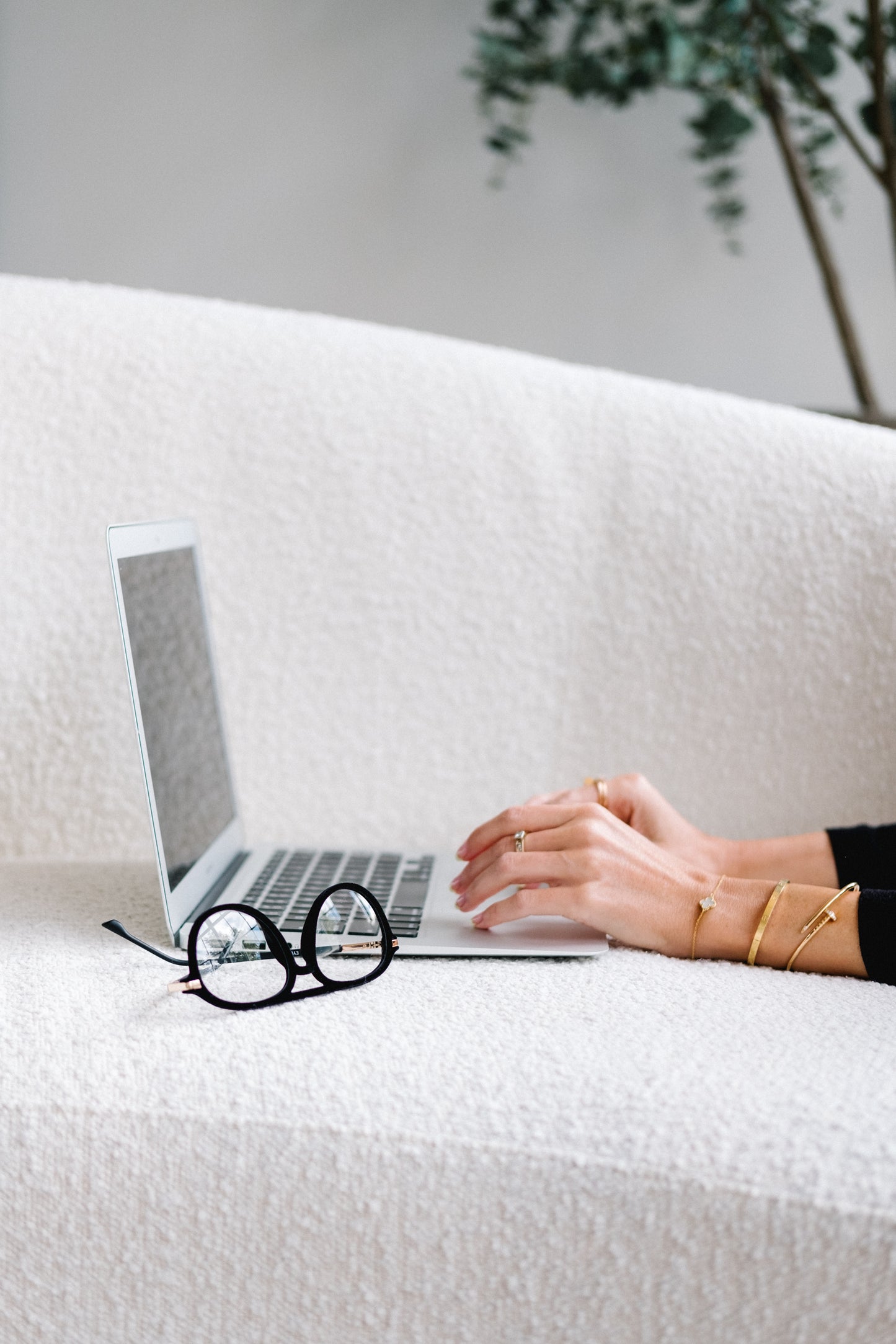 Black velvet Mykonos aviator blue light reading glasses placed next to an open laptop on a textured boucle couch, with a woman’s hands positioned over the keyboard as if she’s about to type. The cozy workspace scene highlights the glasses as a stylish and functional accessory for computer use.
