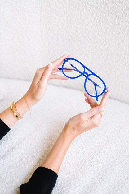Blue velvet Mykonos Aviator reading glasses gently held in a woman’s hands over a soft boucle couch. She’s wearing gold jewelry, and the scene feels chic and refined, highlighting the luxurious velvet texture and elegant blue hue of the frames.