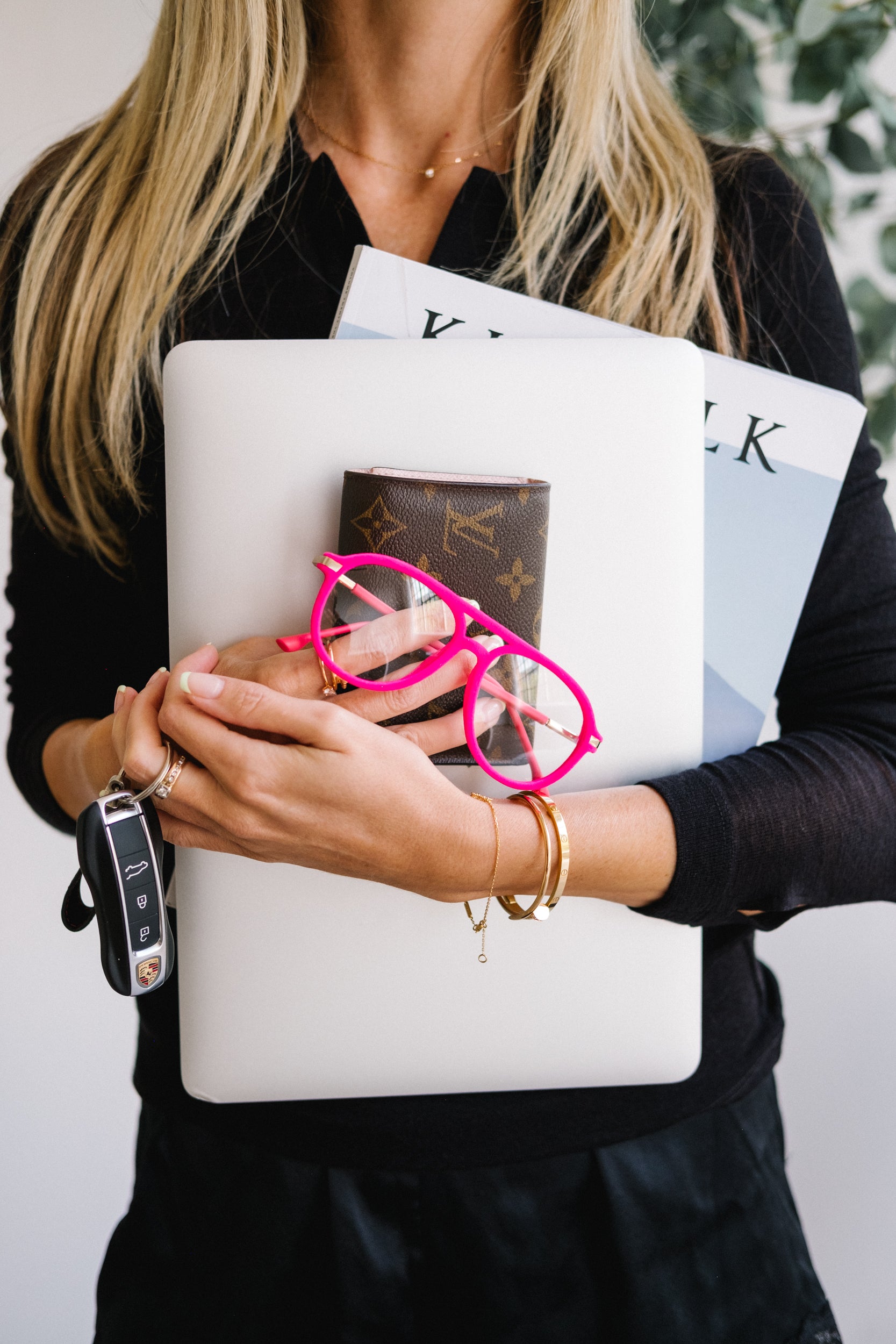 Close-up of a woman’s chest holding pink velvet Mykonos aviator blue light glasses in her hand, along with a laptop, a Kinfolk magazine, Porsche car keys, and a Louis Vuitton wallet. The scene conveys stylish “girl boss” energy, showcasing the glasses as a chic accessory for an on-the-go lifestyle.