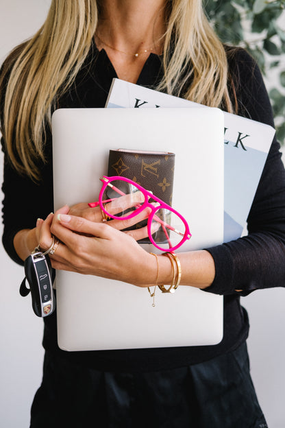 Close-up of a woman’s chest holding pink velvet Mykonos aviator blue light glasses in her hand, along with a laptop, a Kinfolk magazine, Porsche car keys, and a Louis Vuitton wallet. The scene conveys stylish “girl boss” energy, showcasing the glasses as a chic accessory for an on-the-go lifestyle.