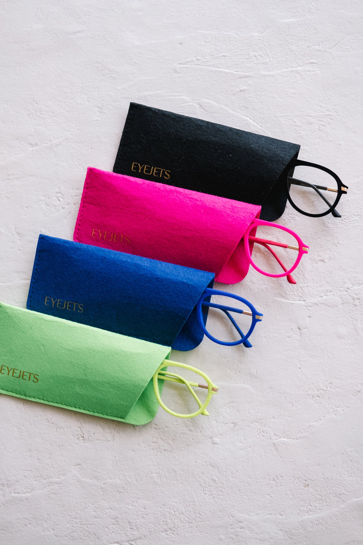 Four Mykonos velvet reading glasses in black, blue, pink, and neon green colors, partially peeking out of their matching pouches on a white desk. The image highlights the soft velvet texture and vibrant color variety of the collection in a clean, modern setting.