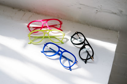 All four velvet Mykonos Aviator reading glasses — black, blue, pink, and neon green — displayed on a white pedestal by a sunlit window. Soft natural light casts gentle shadows, highlighting the luxurious velvet texture and vibrant colors for an elegant, high-end look.