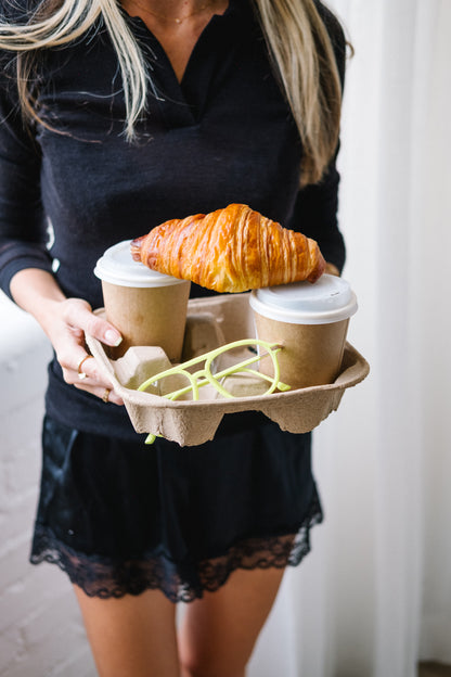 Person wearing a black outfit, holding a coffee tray with two coffees, a croissant, and a pair of neon green velvet aviator glasses. The photo focuses on the stylish details and vibrant pop of the neon frames.