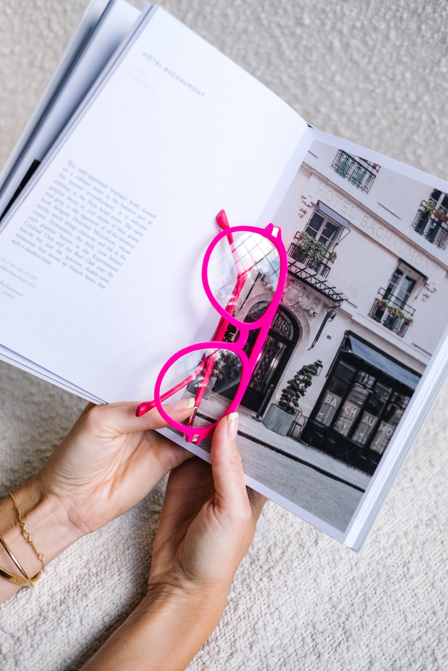 Pink velvet Mykonos aviator blue light glasses resting between the open pages of a Paris City Guide, held by a woman’s hands. The scene evokes a cozy, stylish reading moment, highlighting the glasses as a chic accessory for book lovers and travel enthusiasts.
