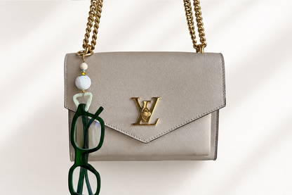 A light beige leather Louis Vuitton shoulder bag with a gold-tone chain strap and a gold-tone LV logo on the front flap. A green eyeglasses keychain charm hangs from the strap.